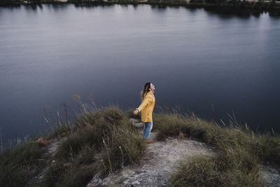 Young woman wearing yellow raincoat standing with arms outstretched at lakeshore