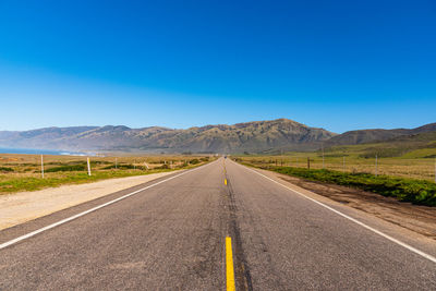 Road leading towards mountains against clear blue sky