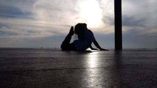 Side view of girl practicing yoga on wooden floor against cloudy sky