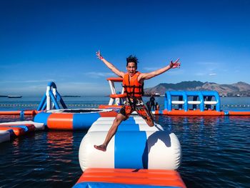 Portrait of happy young man with arms raised jumping on inflatable raft in lake against sky