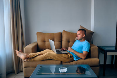 Young adult caucasian man works on laptop lying on sofa. thoughtful man surfing a net lying on couch