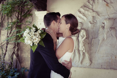 Side view of bridegroom hugging bride against carvings on wall