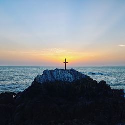 Cross on cliff by sea against sky during sunset