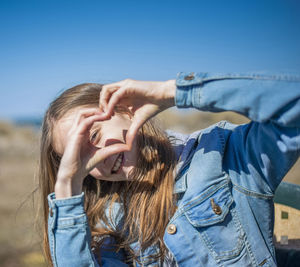 Portrait of girl making heart shape while standing against blue sky