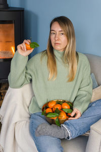 Blonde woman holds a bowl of fresh tangerines while sitting in an armchair by the fireplace.