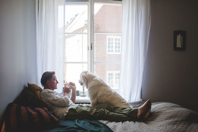 Senior man playing with dog while leaning on bed by window at home