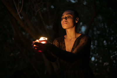 Young woman looking away while standing against trees at night