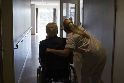 Rear view of female nurse with senior man sitting in wheelchair at hospital corridor