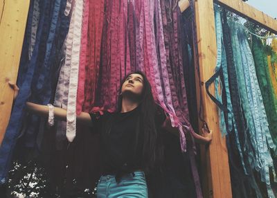 Low angle view of young woman standing by colorful fabrics hanging