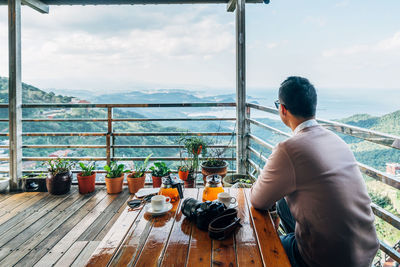 Rear view of man having coffee while sitting at table by mountains against sky