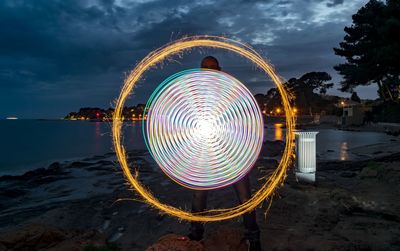 Man spinning illuminated lighting equipment at beach against sky during night