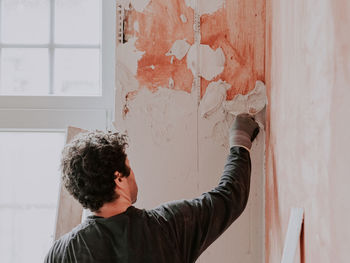 A young caucasian guy applies fresh putty with a small spatula on the wall near the window