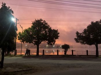 Silhouette trees by street against sky during sunset