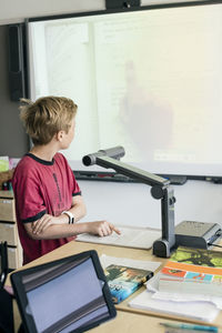 Boy reading document under camera while standing by desk in classroom