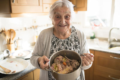 Portrait of smiling senior woman showing a cooking pot of galician stew in the kitchen