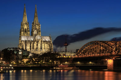 Illuminated cologne cathedral by river against sky at dusk