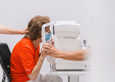 Adult doctor ophthalmologist in protective mask checking eyesight of senior female patient with ophthalmoscope