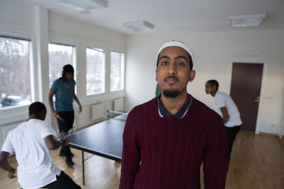 Portrait of man standing while friends playing table tennis in background at games room