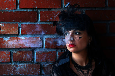 Thoughtful woman in widow hat smoking against brick wall