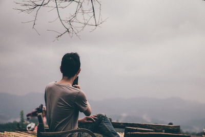 Side view of man photographing with smart phone while sitting on mountain against cloudy sky