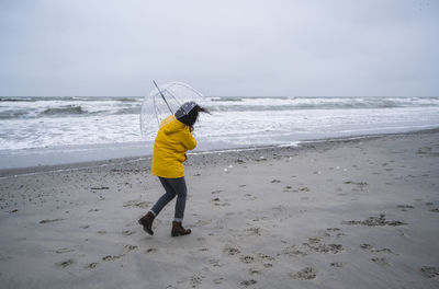 Woman in raincoat walking with umbrella at sea shore against sky