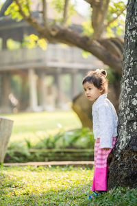 Girl standing on grass by tree trunk against building