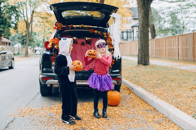 Full length of cute sibling holding pumpkin standing by car