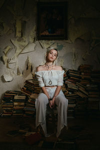 Portrait of young woman sitting on stacked books against weathered wall