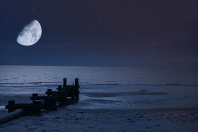 A night sky with a half moon and stars over the beach and ocean with a pipe