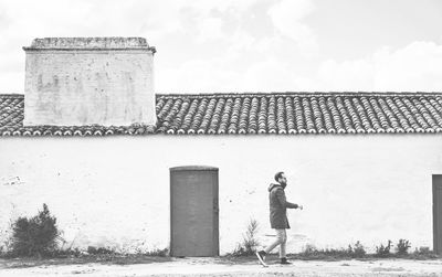 Side view of man walking by house against cloudy sky