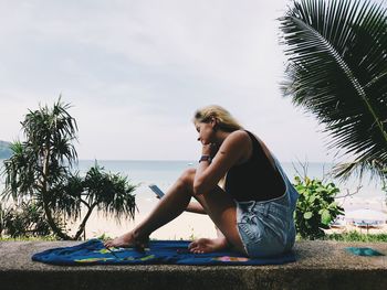 Woman sitting on palm tree by sea against sky