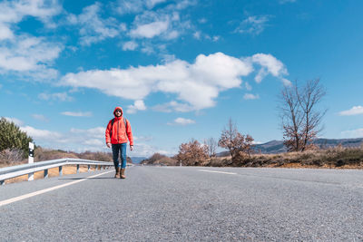 A man wearing a red jacket and jeans walking on the asphalt road under a blue sky. copy space.