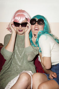 Happy women wearing sunglasses and wigs while sitting on couch against wall