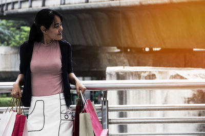 Woman holding shopping bags while standing against railing