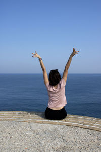 Rear view of happy woman raising arms in front of ocean against clear sky