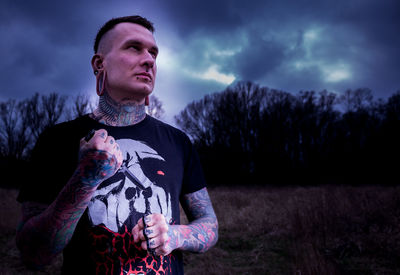 Tattooed man holding knife while standing on field against sky at dusk