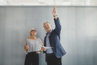 Mature businessman holding documents while discussing with businesswoman against wall in new office