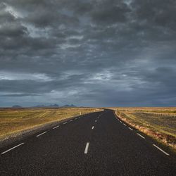 Road passing through field against storm clouds