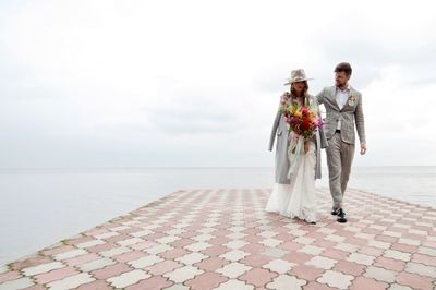 Newly married couple walking on pier over lake against cloudy sky