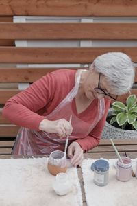 Senior gray haired woman with glasses covering with white glaze handmade clay vase by brush