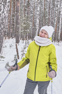 Smiling senior woman in yellow jacket, white knitted hat, scarf with ski poles in   snowy forest