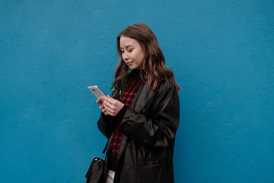 Young woman using phone while standing against blue wall