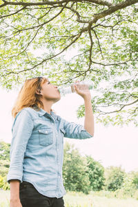 Woman drinking water from bottle while standing against trees