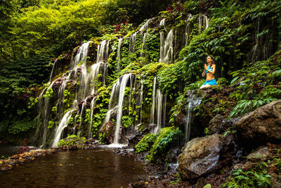 Full length of woman sitting by waterfall in forest