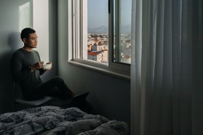 Side view of young woman looking through window at home