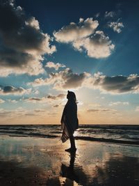 Silhouette person wrapped in blanket standing at beach during sunset