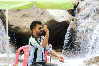 Young man sitting on chair against waterfall