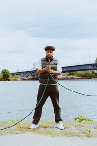 Full length portrait of confident young man holding garden hose near river