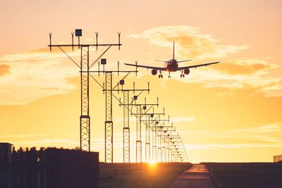 Low angle view of airplane flying by electricity pylon against sky during sunset