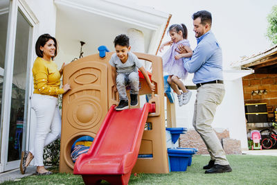 Full body of joyful little ethnic girl playing on slide while spending time in backyard with young parents on sunny day